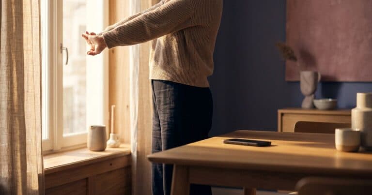 Person streckt sich am Fenster, das Smartphone liegt weiter hinten auf dem Tisch